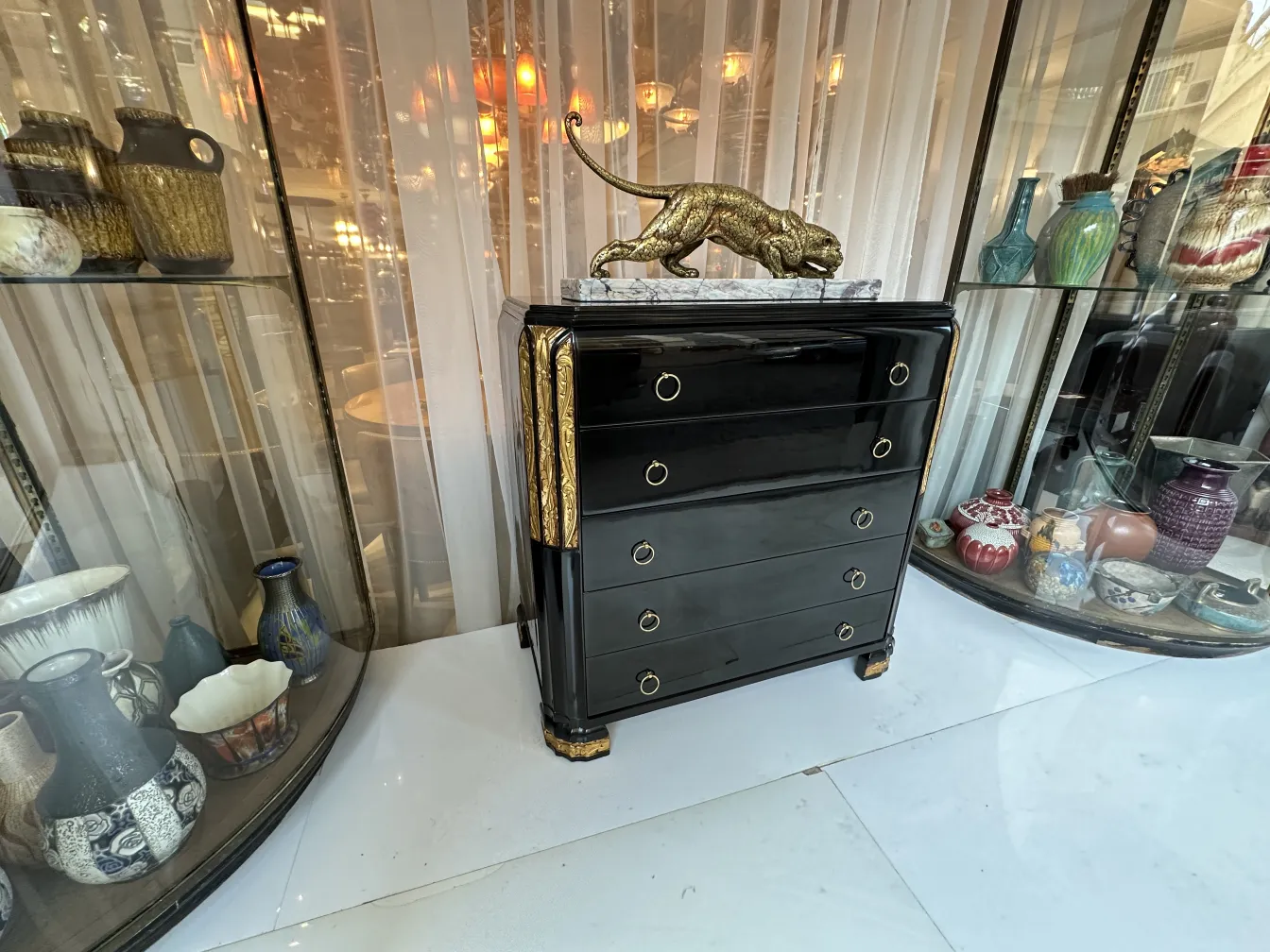 Black antique cabinet with golden details and a leopard decoration on top. Background features antique vases.