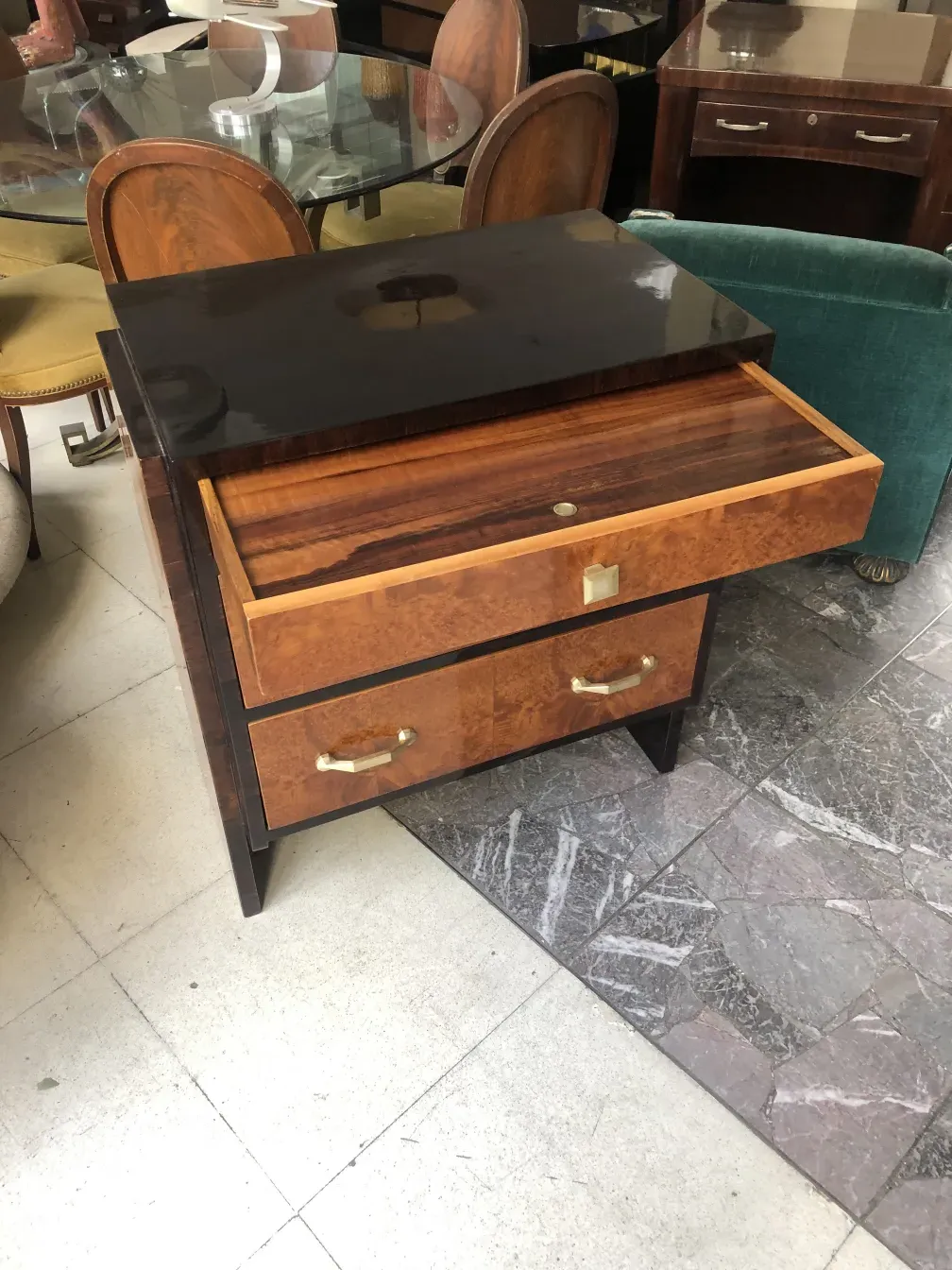 Cassettiera antica in legno con due cassetti e superficie lucida, circondata da sedie e tavoli.