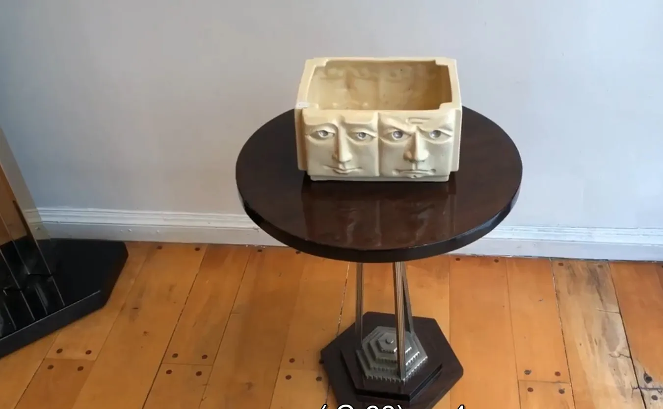 Contêiner retangular de cerâmica antiga com rostos esculpidos, colocado sobre uma mesa escura.