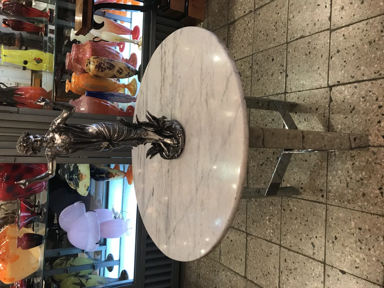 Sculpture antique de figure féminine sur une table ronde, avec des vases colorés en arrière-plan.