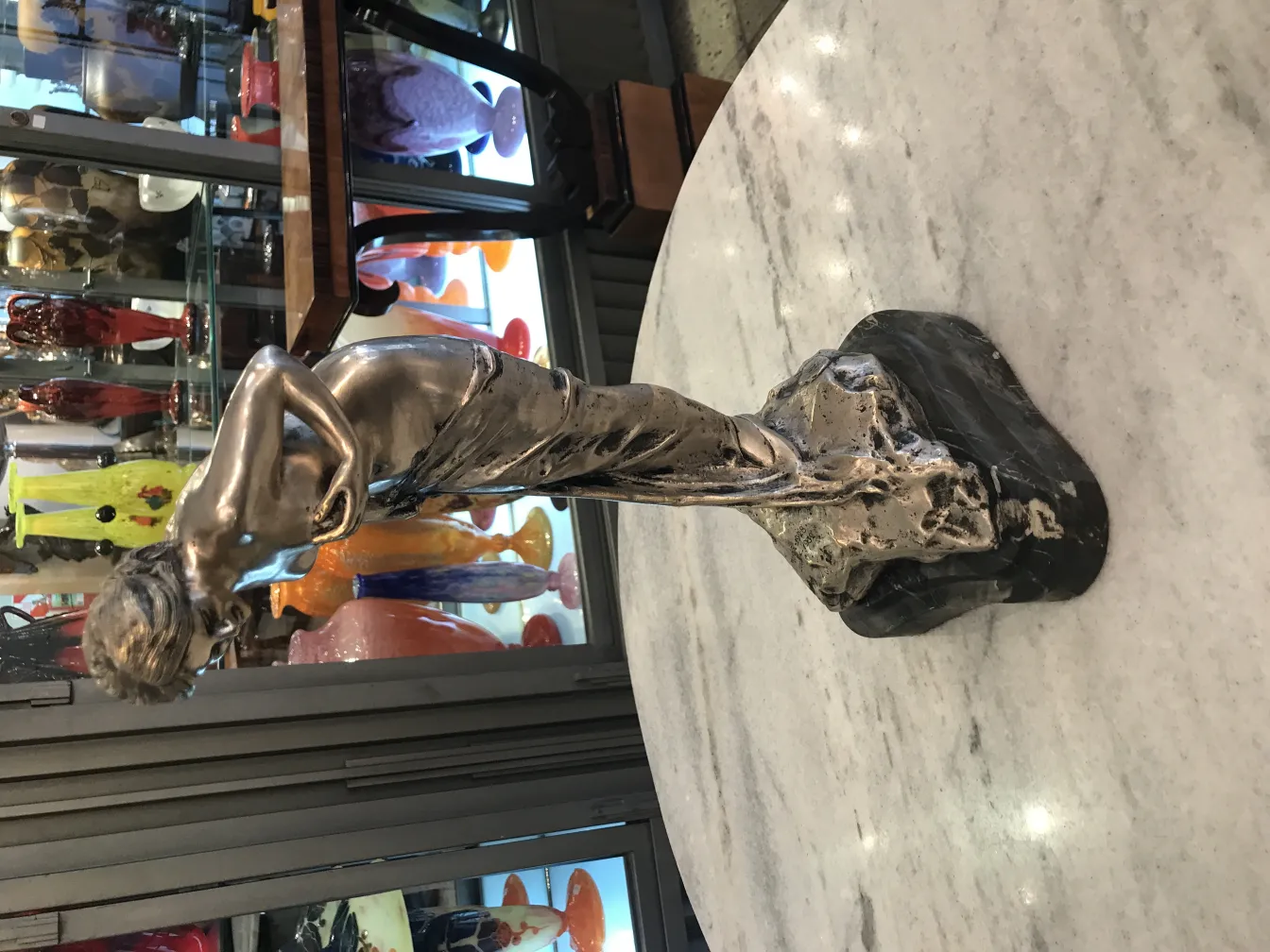 Antike Skulptur einer eleganten Figur mit dunklem Sockel, umgeben von bunten Objekten.