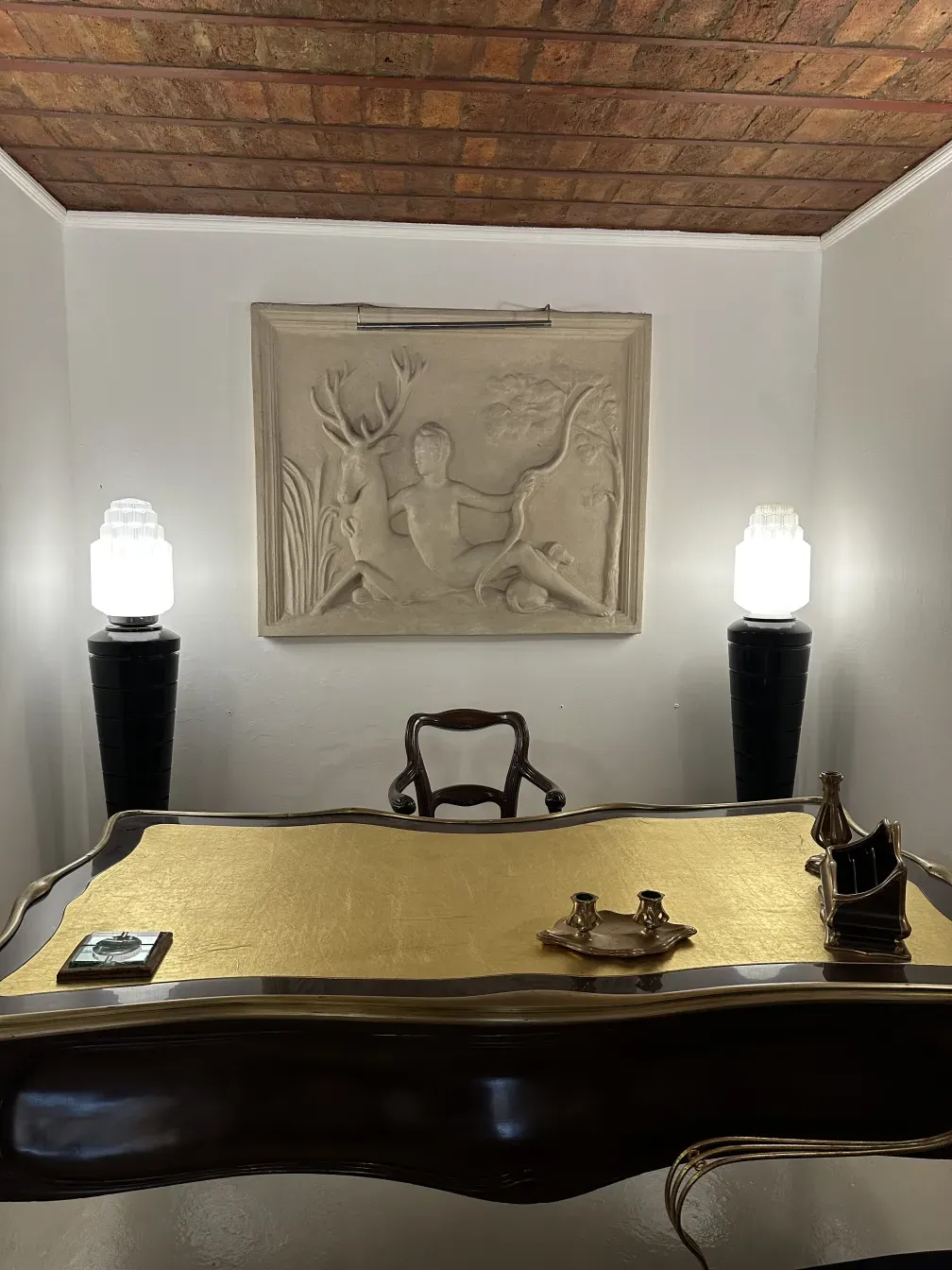 Bureau ancien en bois avec détails dorés. En arrière-plan, une œuvre d'art en relief et un mur en briques.