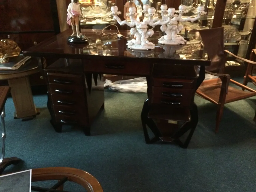 Mesa antiga com acabamentos escuros e desenhos decorativos. Cercada por itens antigos.