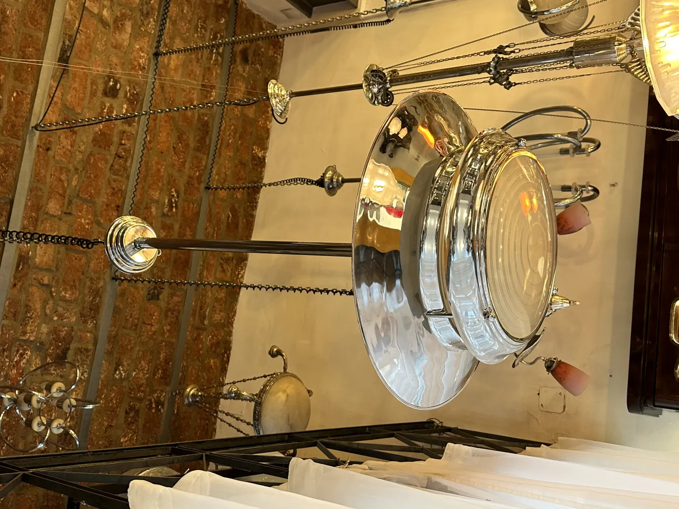 Lampada a sospensione antica con un design lucido, circondata da catene e sfondo di mattoni a vista.