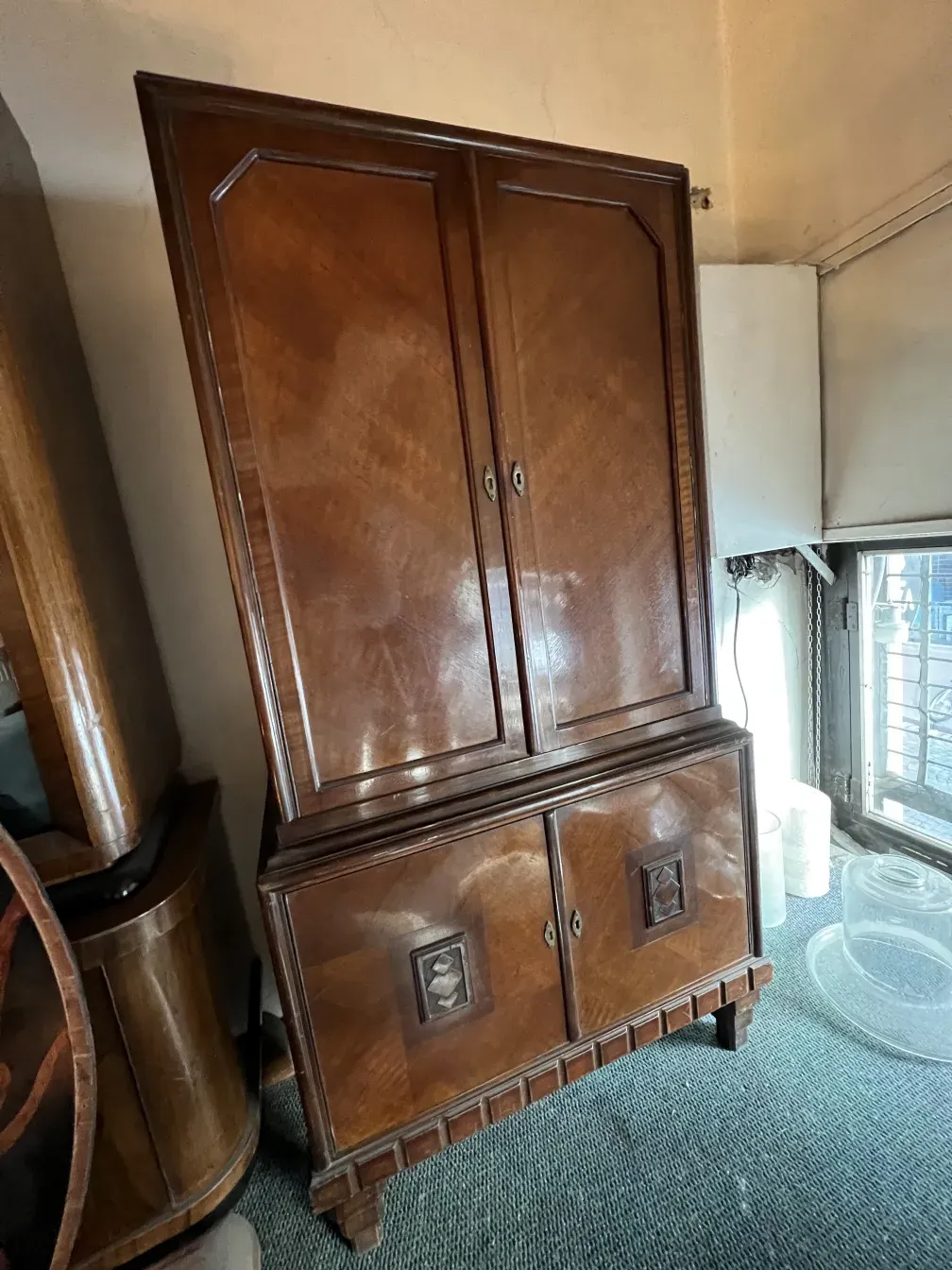 Biblioteca antigua de madera con diseño detallado y puertas dobles, rodeada de otros muebles de madera en un ambiente acogedor.