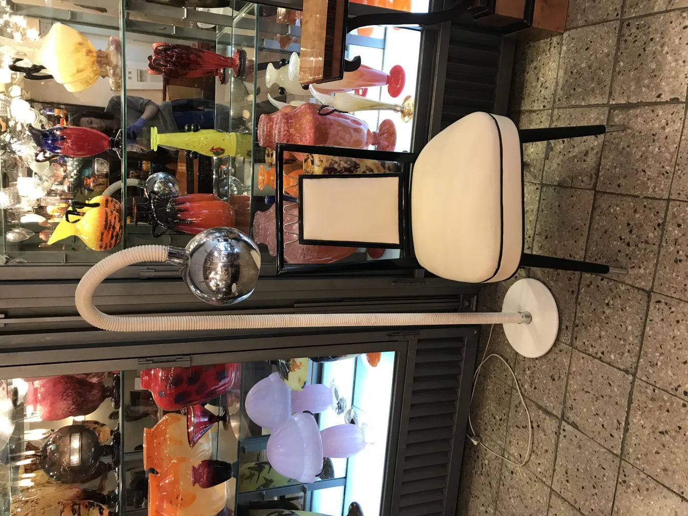 Luminária de pé antiga com base branca e detalhes cromados, cercada por antiguidades coloridas.