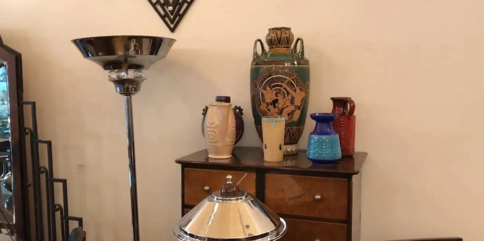 Luminária de pé antiga com base de metal e cúpula clássica, rodeada de itens decorativos coloridos.