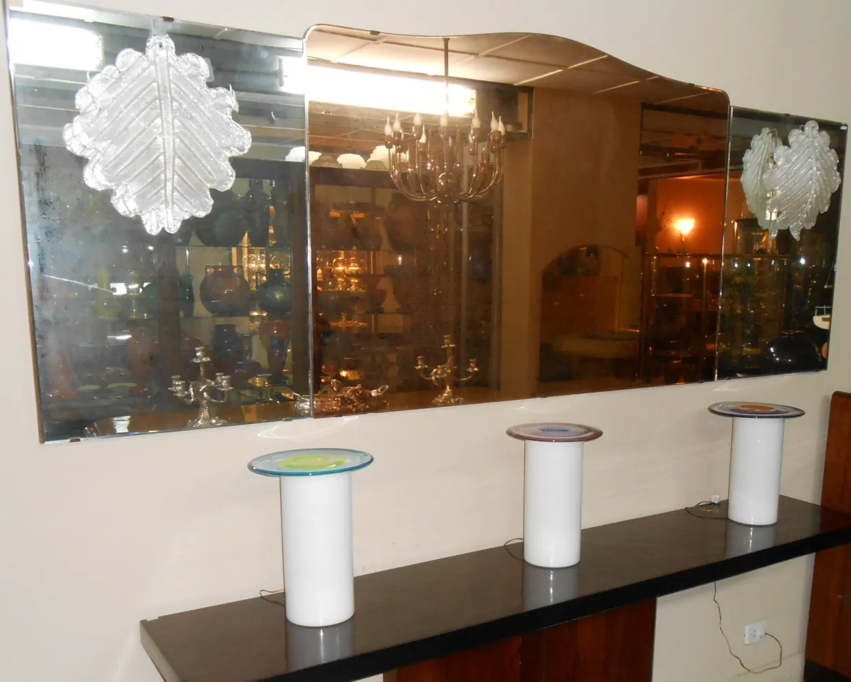 Miroir ancien avec des décorations en forme de feuille, reflétant l'environnement. Trois piédestaux blancs autour.