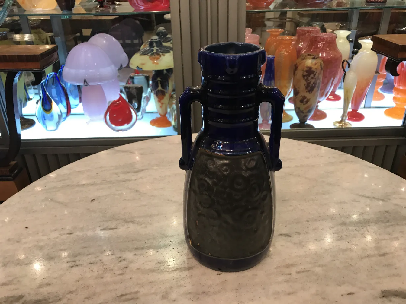 Jarra antigua de cerámica azul y negra con diseño en espiral, en un entorno lleno de antigüedades coloridas.