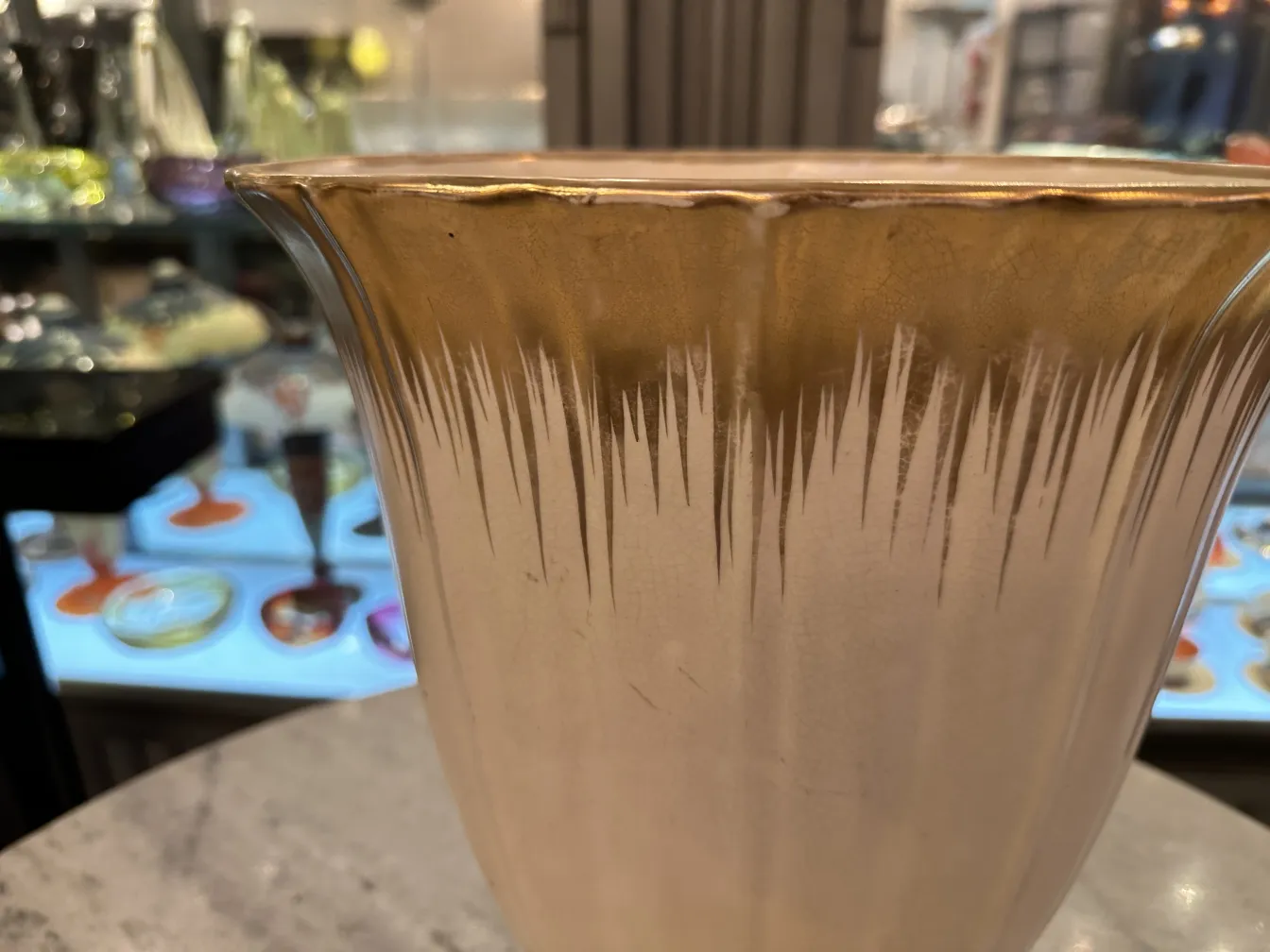 Vaso de cerâmica antiga com decorações douradas e design elegante, fundo desfocado com objetos variados.