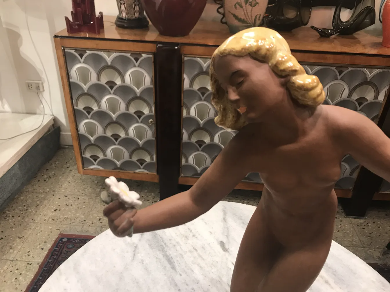 Figura di ceramica antica di una giovane donna che sorride, con una fiore e uno sfondo di arredamento classico.