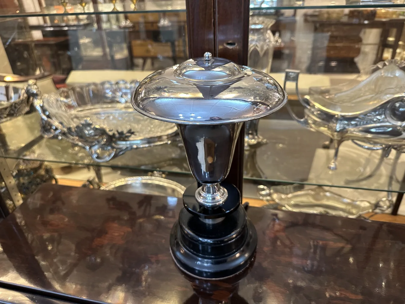 Luminária de mesa antiga com base preta brilhante e tampa prateada em uma vitrine decorativa.