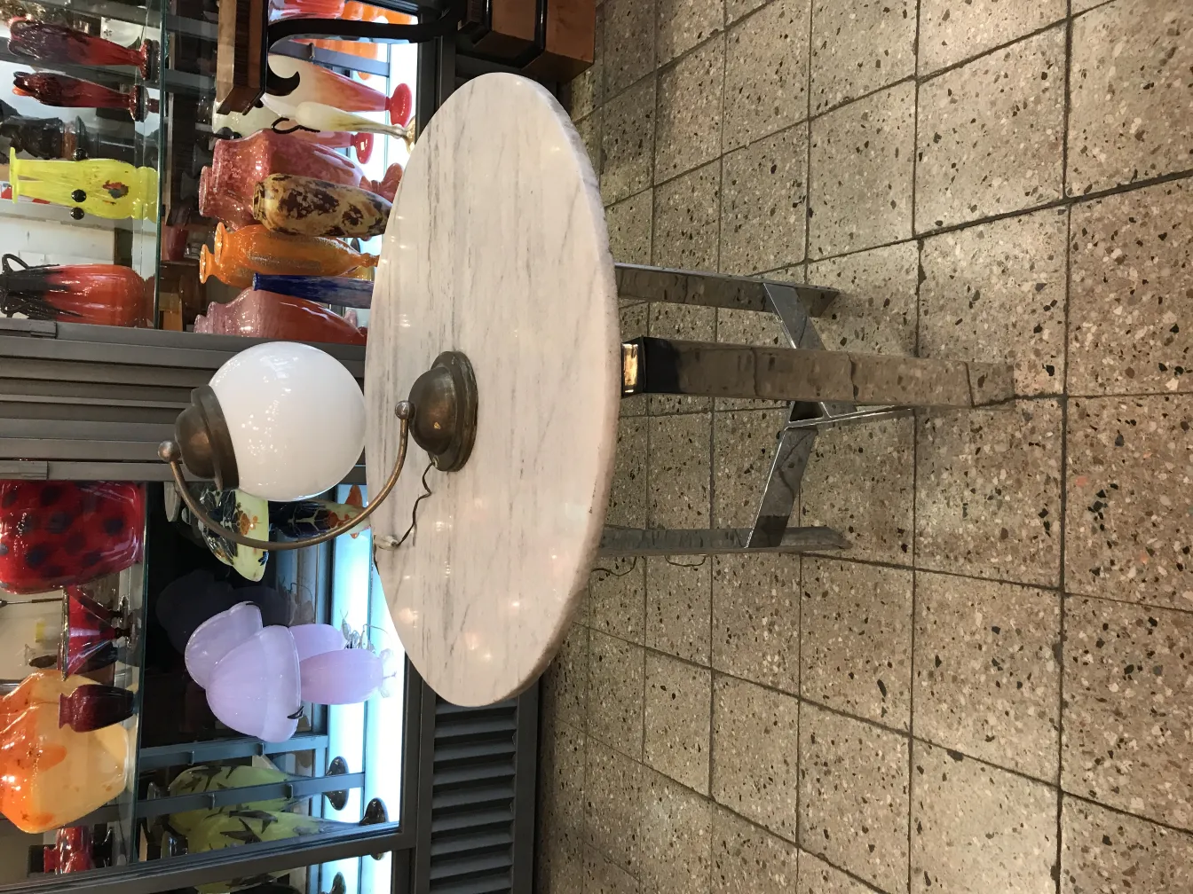 Antigua lámpara de mesa con base metálica y pantalla blanca, rodeada de objetos decorativos coloridos.