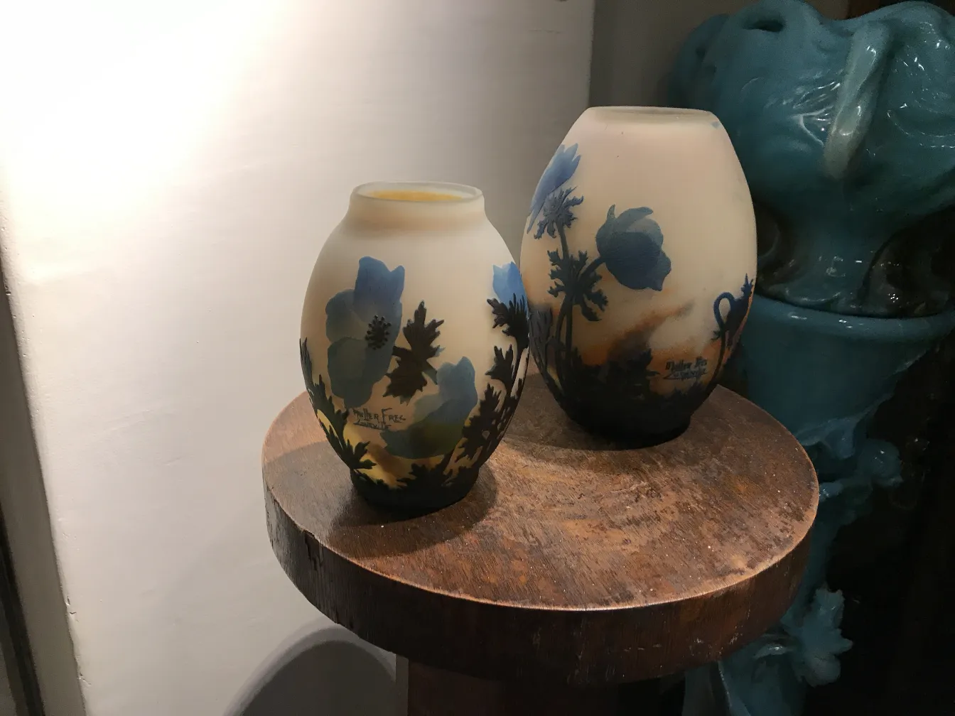 Dois vasos de vidro assinados por Muller Fres com flores azuis e folhas em um ambiente antigo.