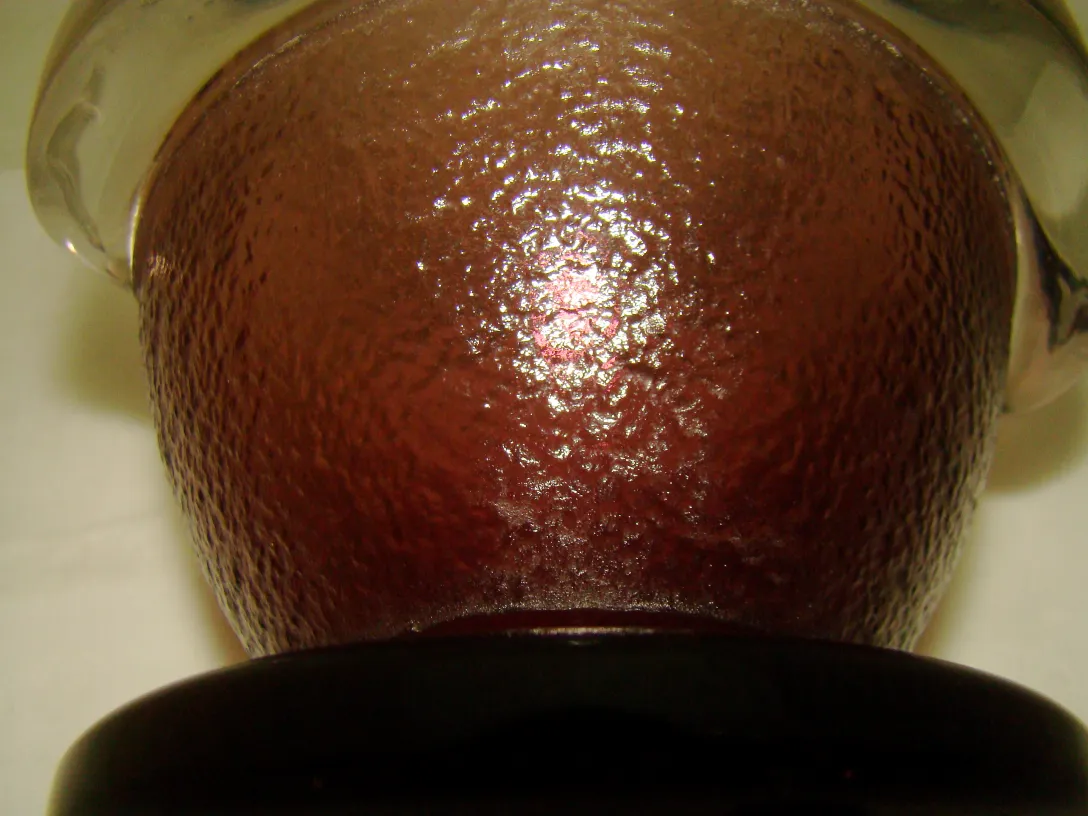 Vidro antigo de tonalidade avermelhada com textura e assinatura na base. Ambiente suave e claro.