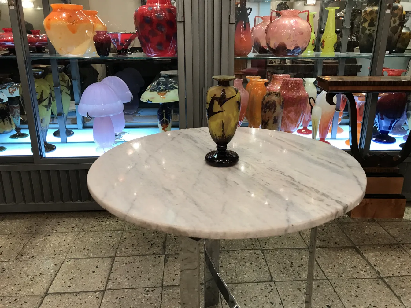 Antigua pieza de vidrio firmado Le Verre en una mesa blanca con otros objetos coloridos al fondo.