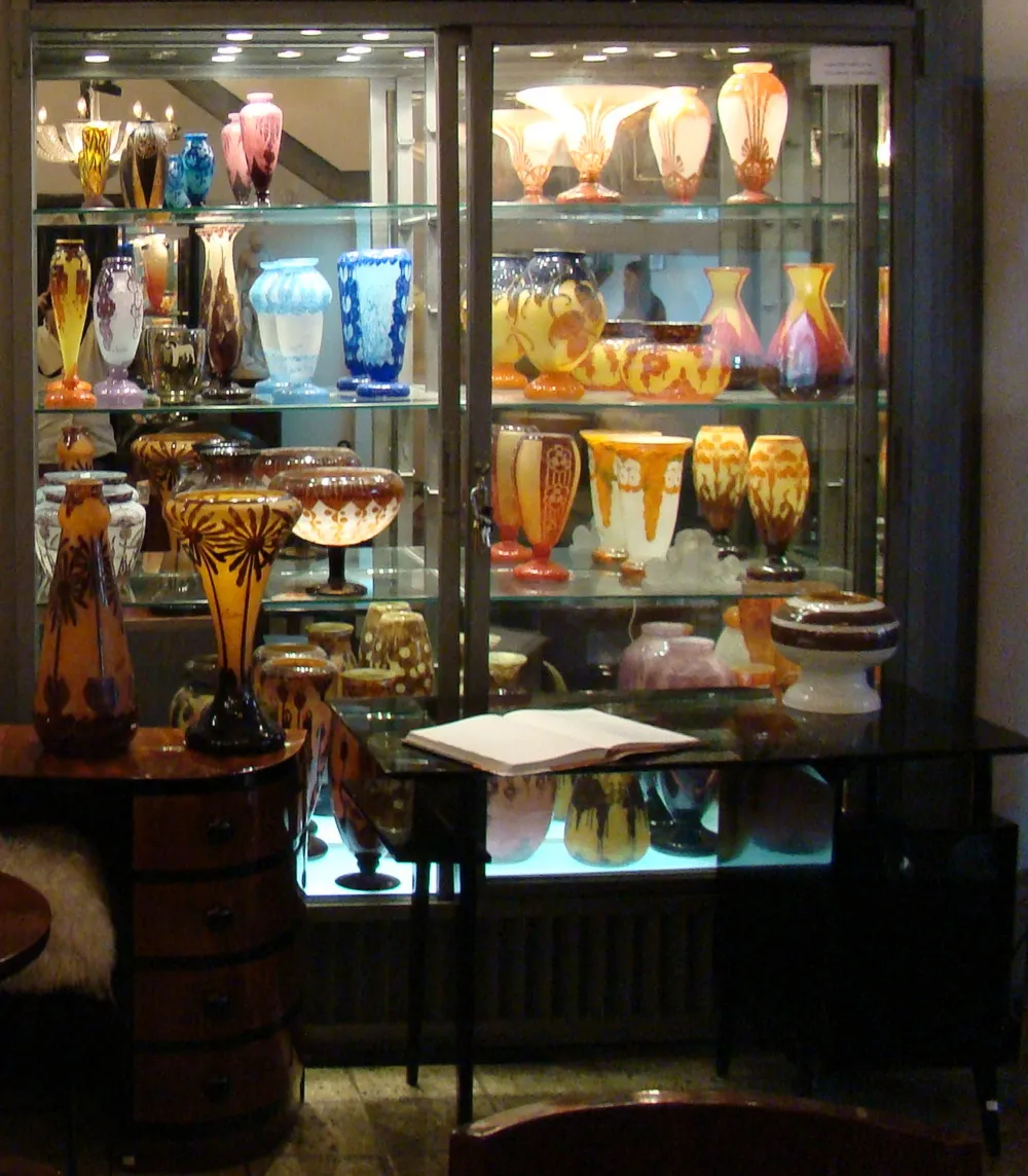 Signiertes Glas von Le Verre in einer Vitrine präsentiert, Antike mit verschiedenen Farben im Hintergrund.