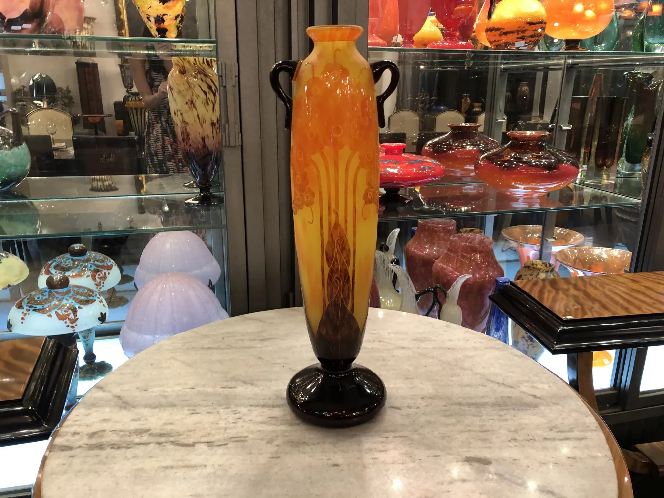 Vaso de vidro assinado Le Verre com detalhes decorativos em laranja e amarelo em um fundo variado.