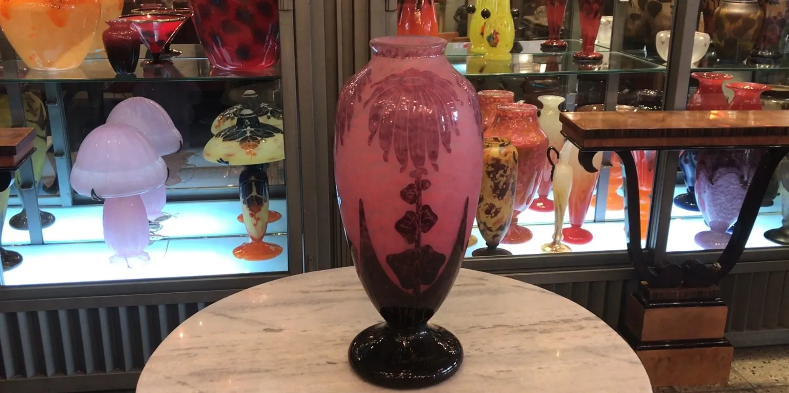 Antique glass vase with floral design in pink, set against a display case filled with other glass items.