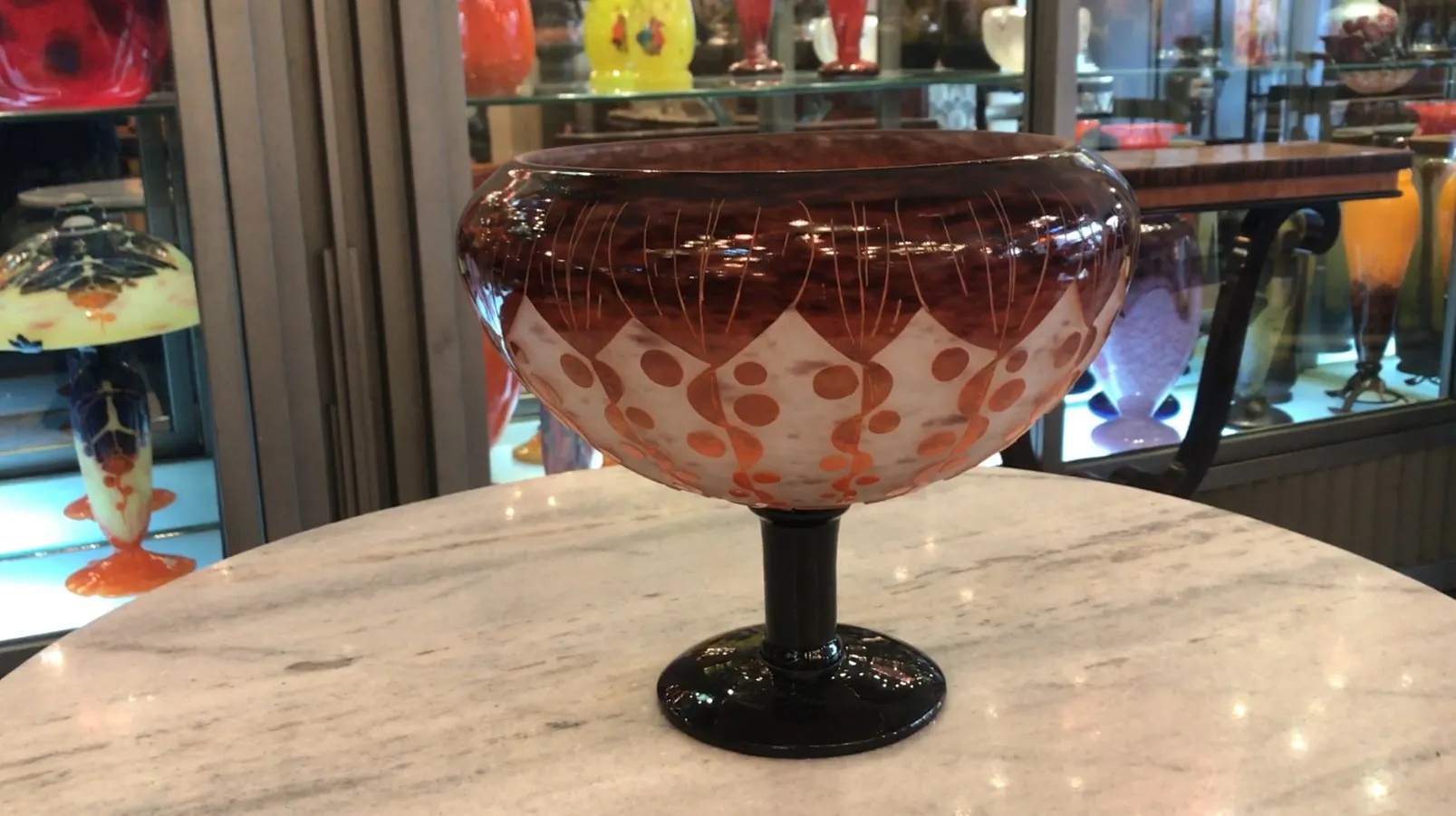 Vidro assinado Le Verre com desenho decorativo em tons quentes, em ambiente de antiguidades.