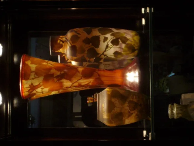 Três vasos antigos assinados da Daum Nancy em cores quentes, exibidos em uma vitrine. Ambiente elegante.