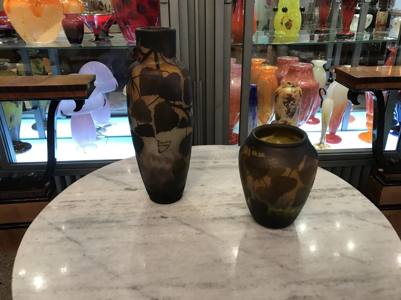 两件达穆·南西签名的古董玻璃置于大理石桌上，背景色彩斑斓的其他古董。