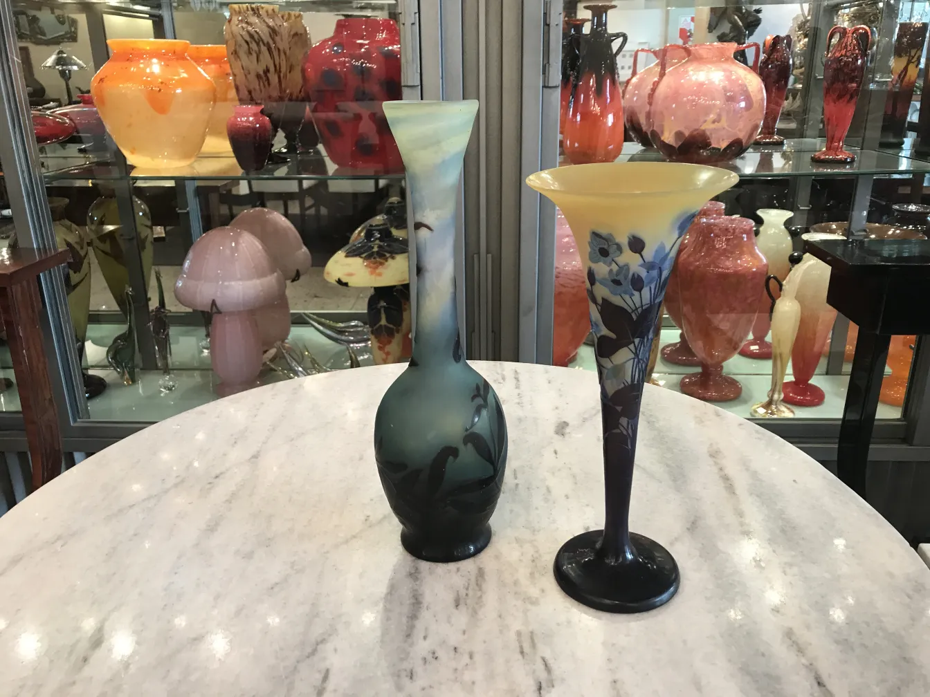 Dois vasos de vidro antigos com decoração floral, um em tons escuros e outro em cores claras, cercados por outros objetos antigos.