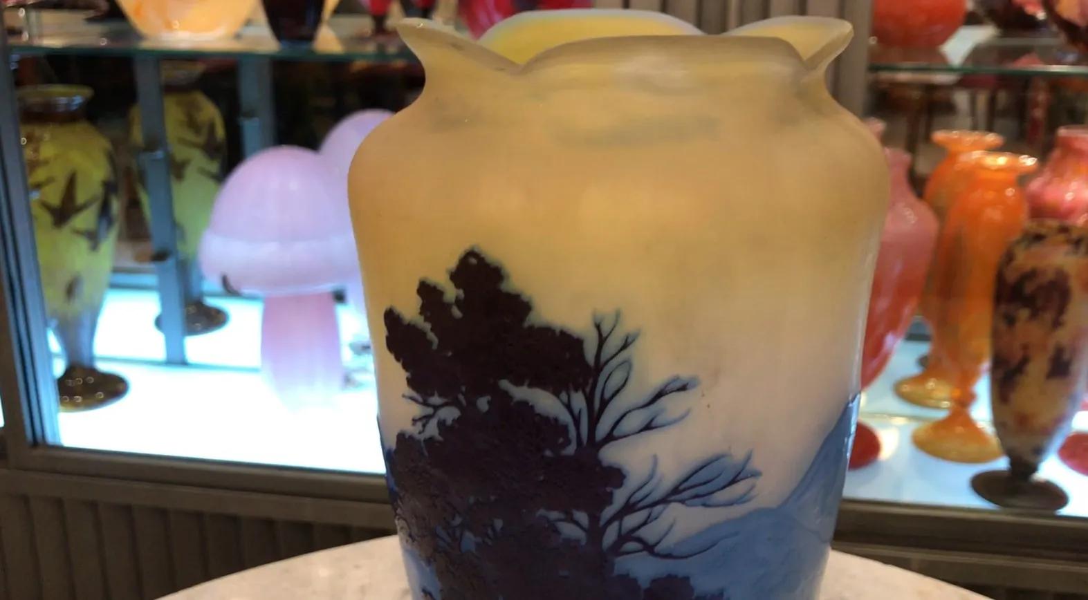 Vaso antigo com design naturalista em tons amarelos e azuis, assinado Gallé, cercado por outros itens decorativos.