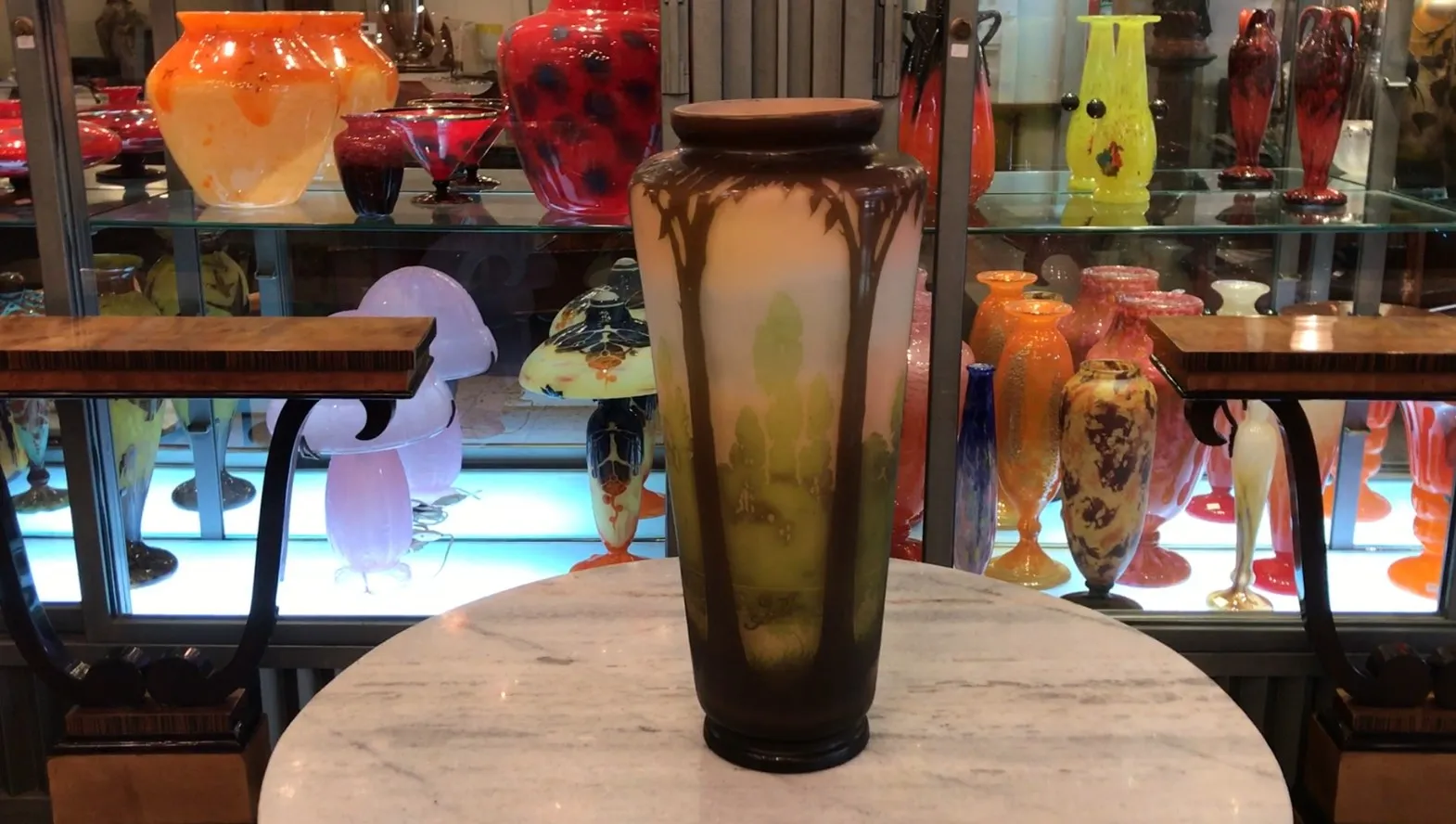 Vaso antigo de vidro assinado Gallé com decoração natural, cercado por outras antiguidades coloridas em uma vitrine.