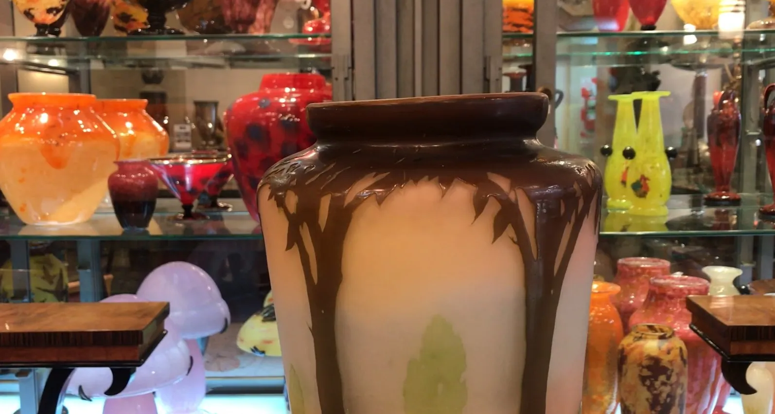 Antica giara in vetro firmato Gallé, decorazioni in tonalità marroni e verdi su sfondo chiaro. Circondata da altri oggetti decorativi.