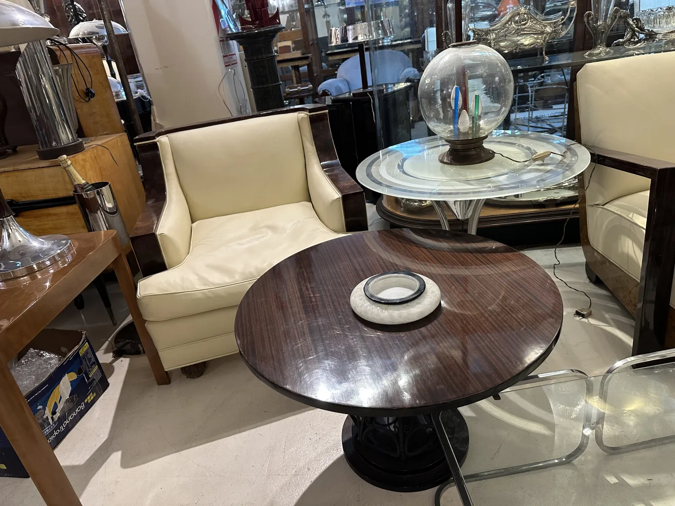 tavolino antico con piano rotondo e base decorativa, in un ambiente elegante con sedie chiare.