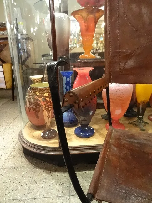 2 poltronas antigas ao lado de uma vitrine com coloridos vasos de vidro.