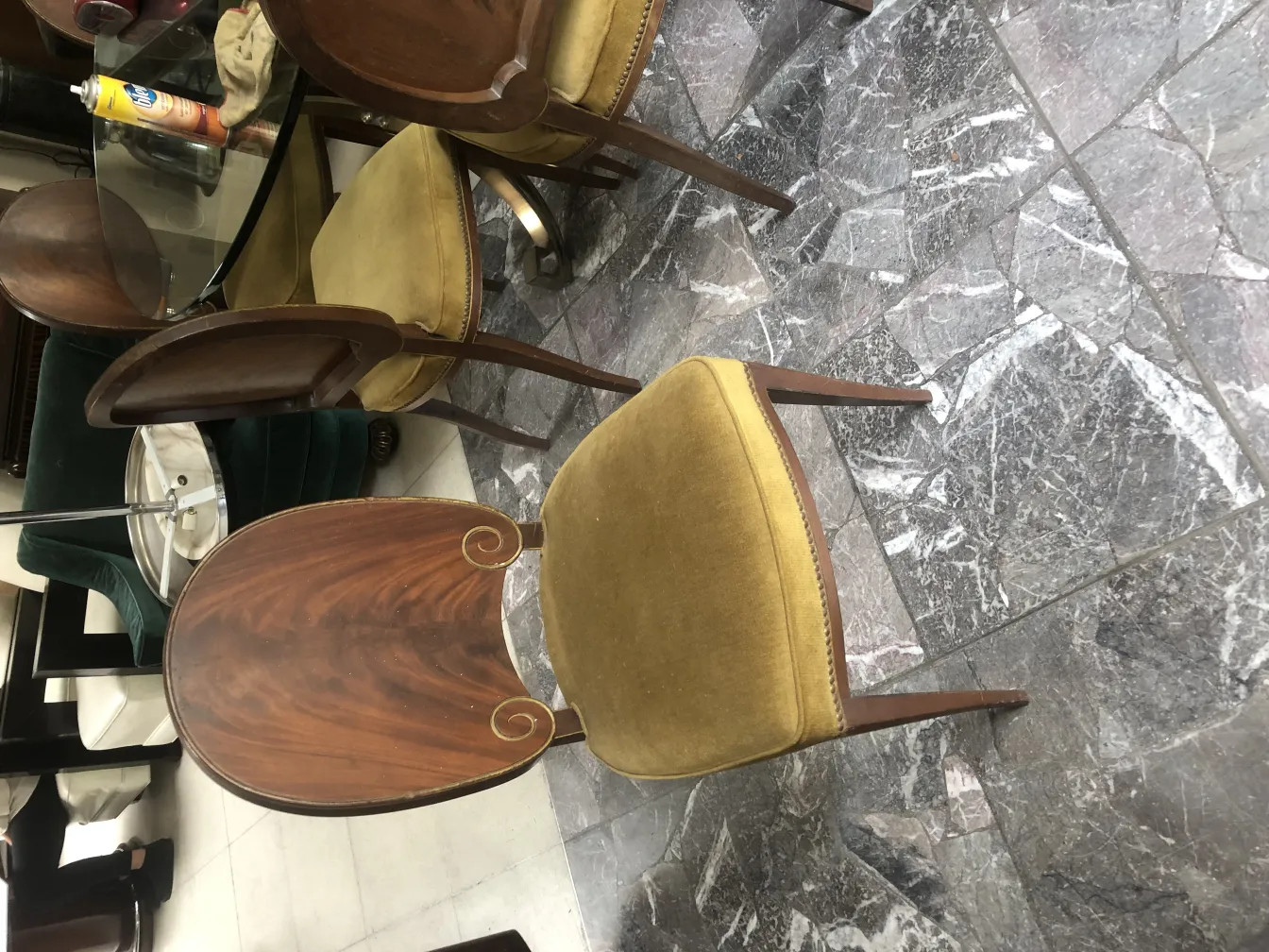 Vista de 12 sillas antiguas con tela verde y un diseño elegante en el respaldo en un ambiente que incluye una mesa de vidrio.