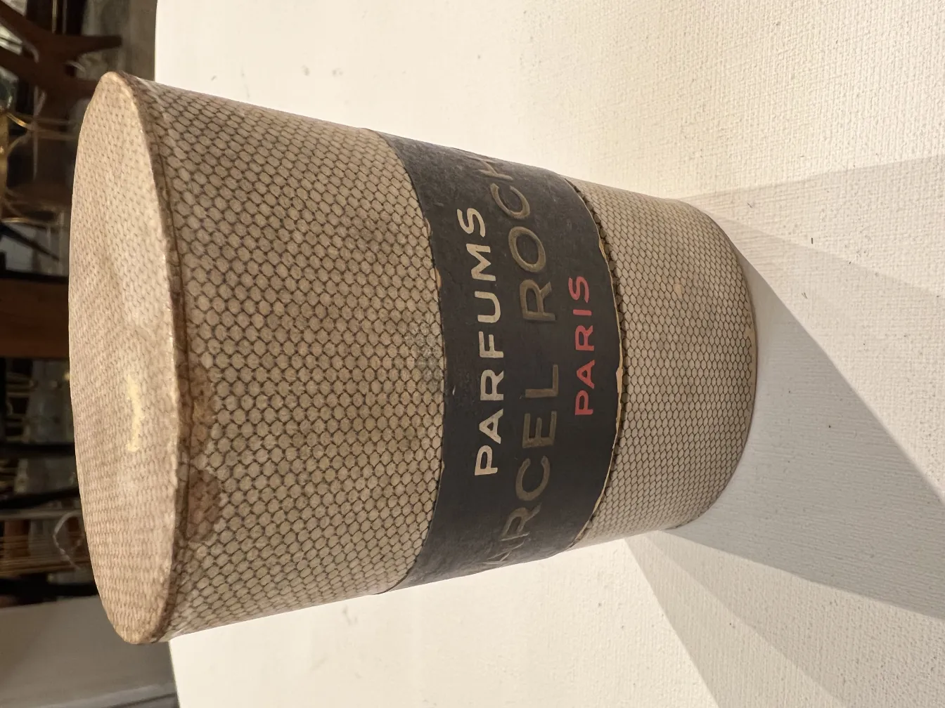 Frasco de perfume antiguo de forma cilíndrica con etiqueta de París, fondo claro.