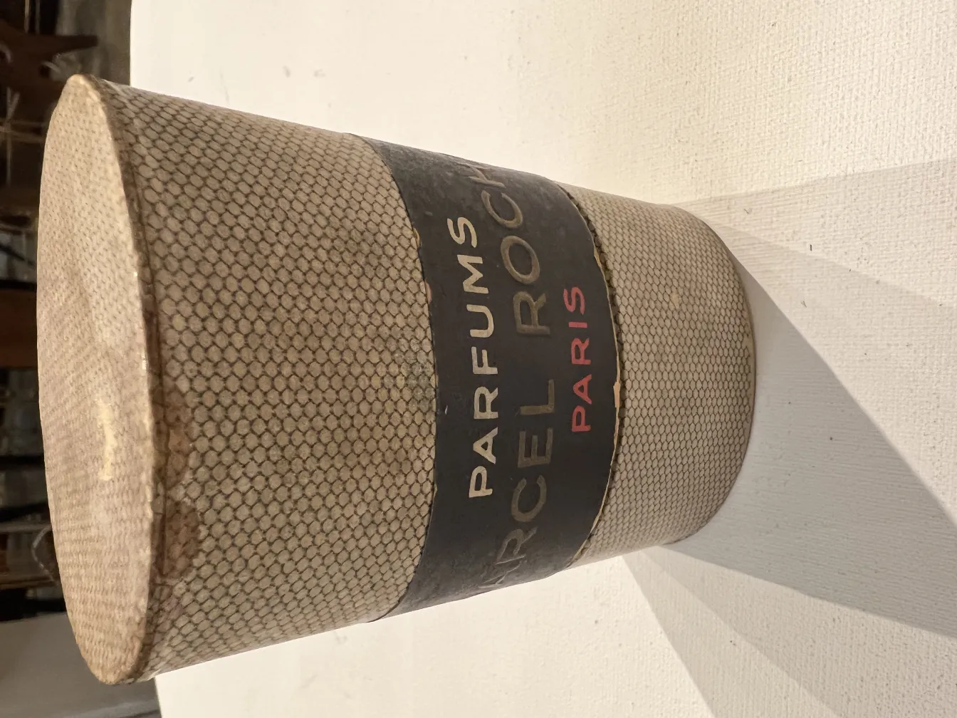 Frasco de perfume antigo em forma cilíndrica com detalhes decorativos, fundo claro.