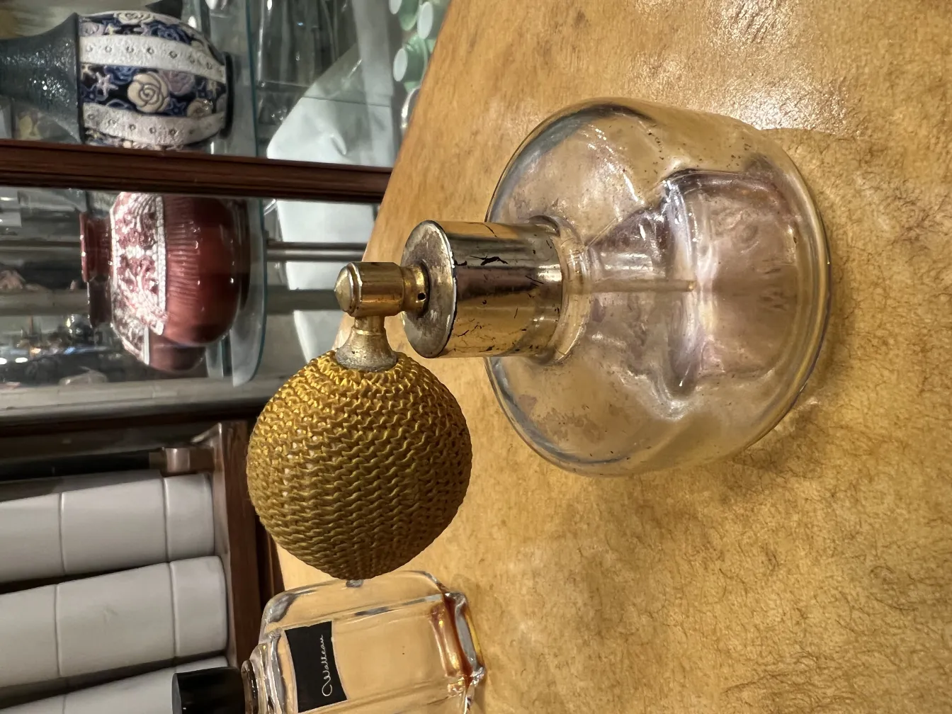 frasco de perfume antiguo con atomizador esférico dorado y base clara, fondo desenfocado de objetos diversos