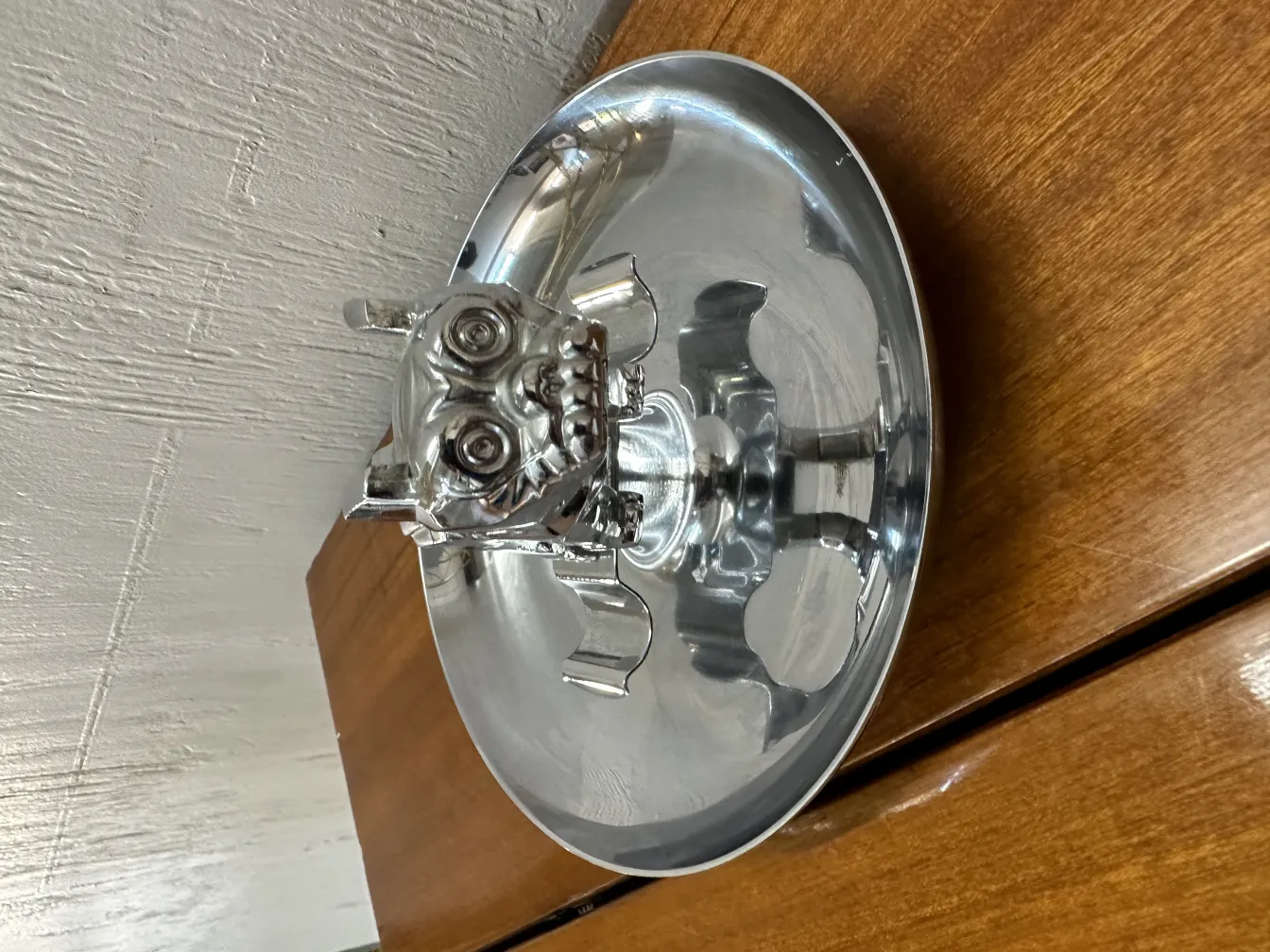 Cenicero antiguo con figura de animal en metal brillante, sobre superficie de madera