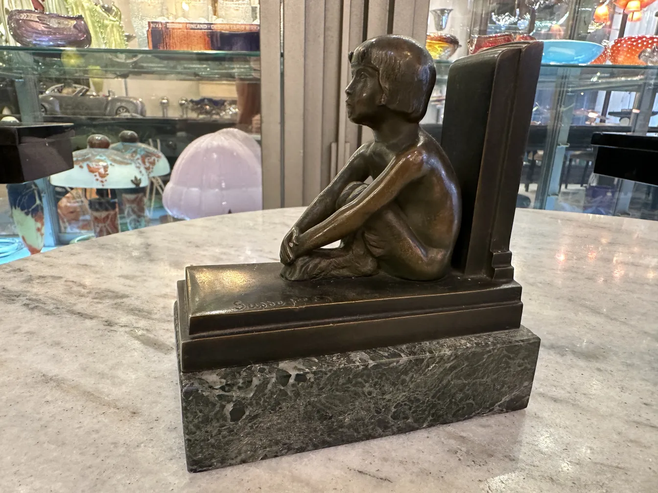 Scultura antica di bambino seduto come fermalibri su marmo, sfondo vario.