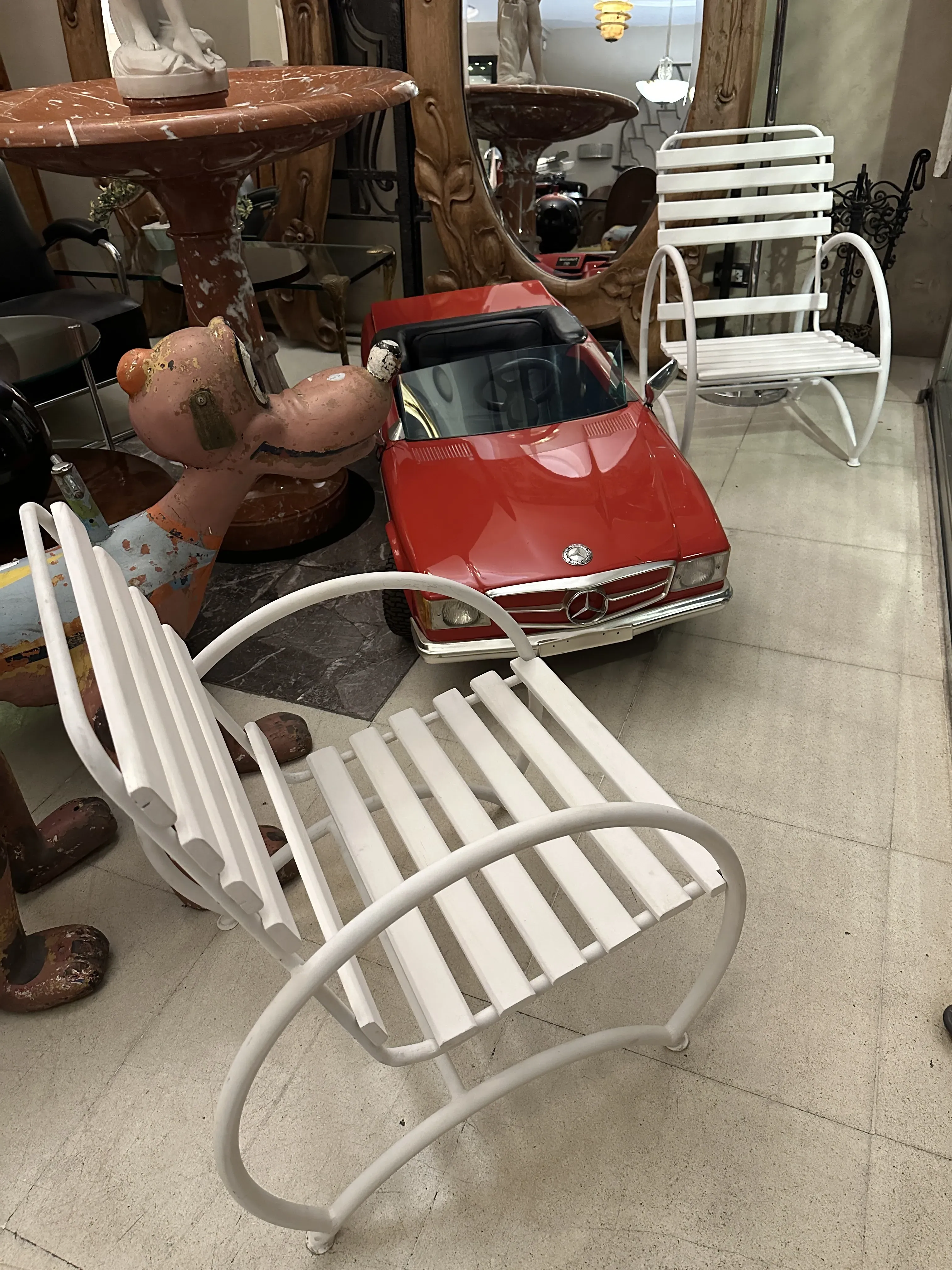 Deux fauteuils de jardin anciens au design classique dans un cadre décoratif avec d'autres objets et une voiture rouge.