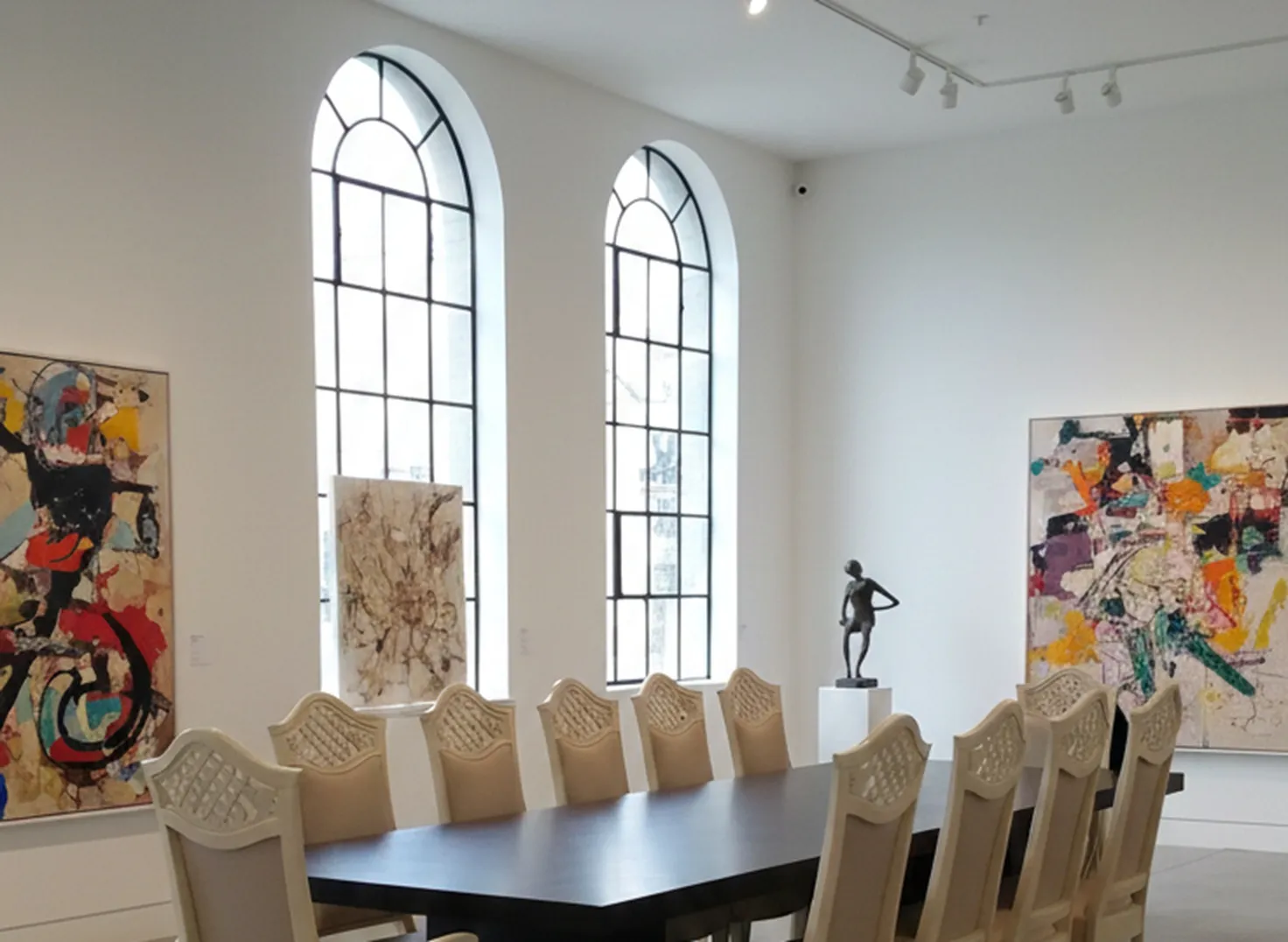 Vista de 12 cadeiras antigas dispostas em uma mesa ampla dentro de uma sala clara com arte contemporânea