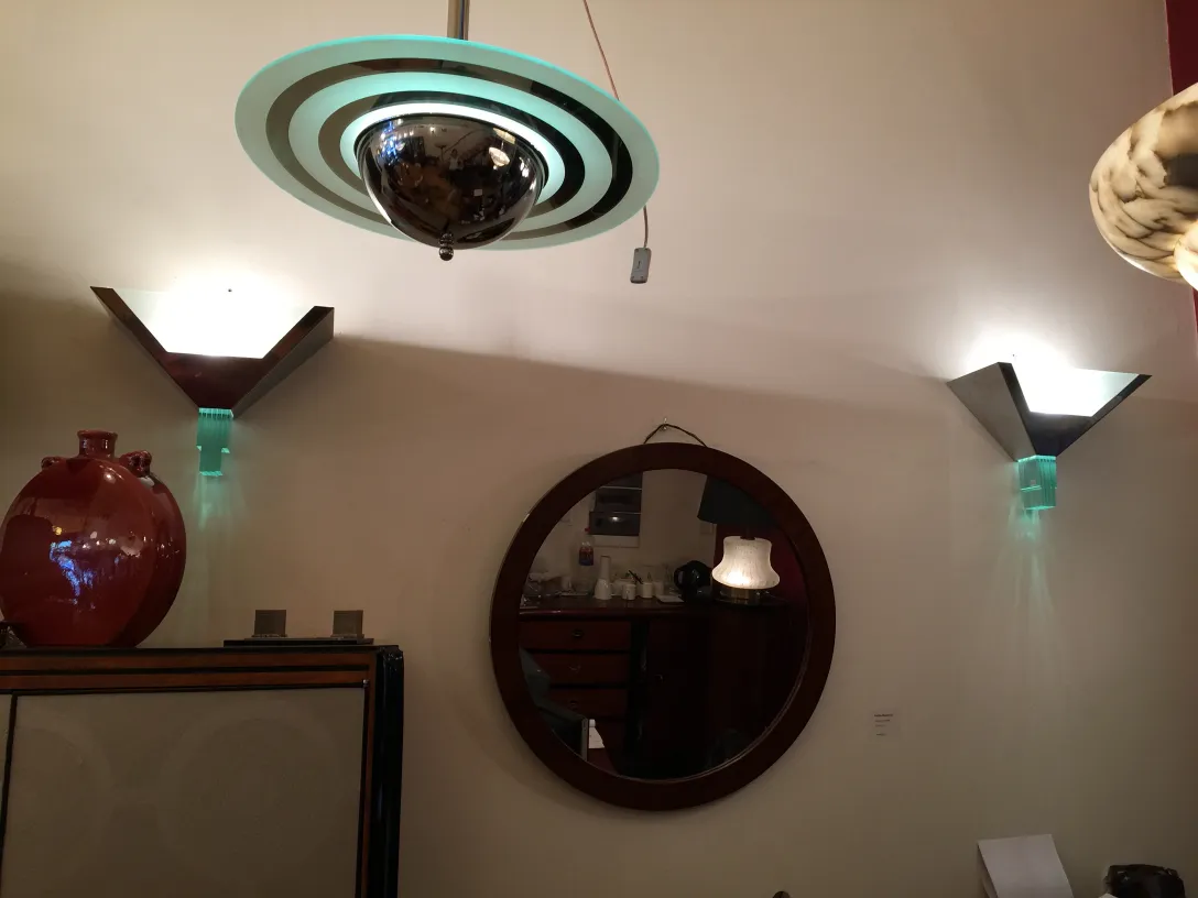 Paire d'appliques anciennes éclairant un mur, à côté d'un vase rouge et d'un miroir rond.